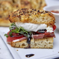 Herbaceous crunchy no-knead focaccia topped with prosciutto, tomatoes, balsamic glaze, microgreens, creamy burrata, olive oil & sea salt flakes making it one of the best sandwiches you'll ever sink your teeth into! Indulge in a Scrumptious Prosciutto, Tomato, & Burrata Focaccia Sandwich, no knead focaccia, Italian sandwich, burrata sandwich, crusty bread